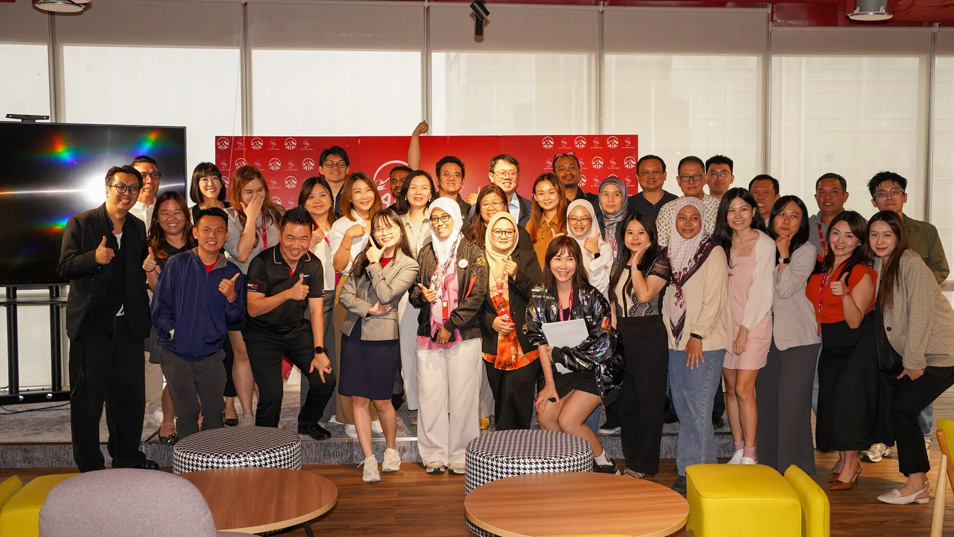 A large group of advisors and trainers smiling and giving thumbs up in front of a red backdrop at the end of a full-day AI training in Johor Bahru, 14 October 2026